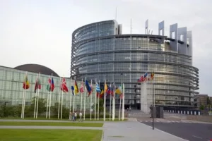 L'esterno della sede del Parlamento europeo a Strasburgo, un moderno edificio di pianta circolare con struttura in vetro e acciaio. Antistanti, la teoria delle bandiere nazionali dei Paesi membri e il viale d'accesso.